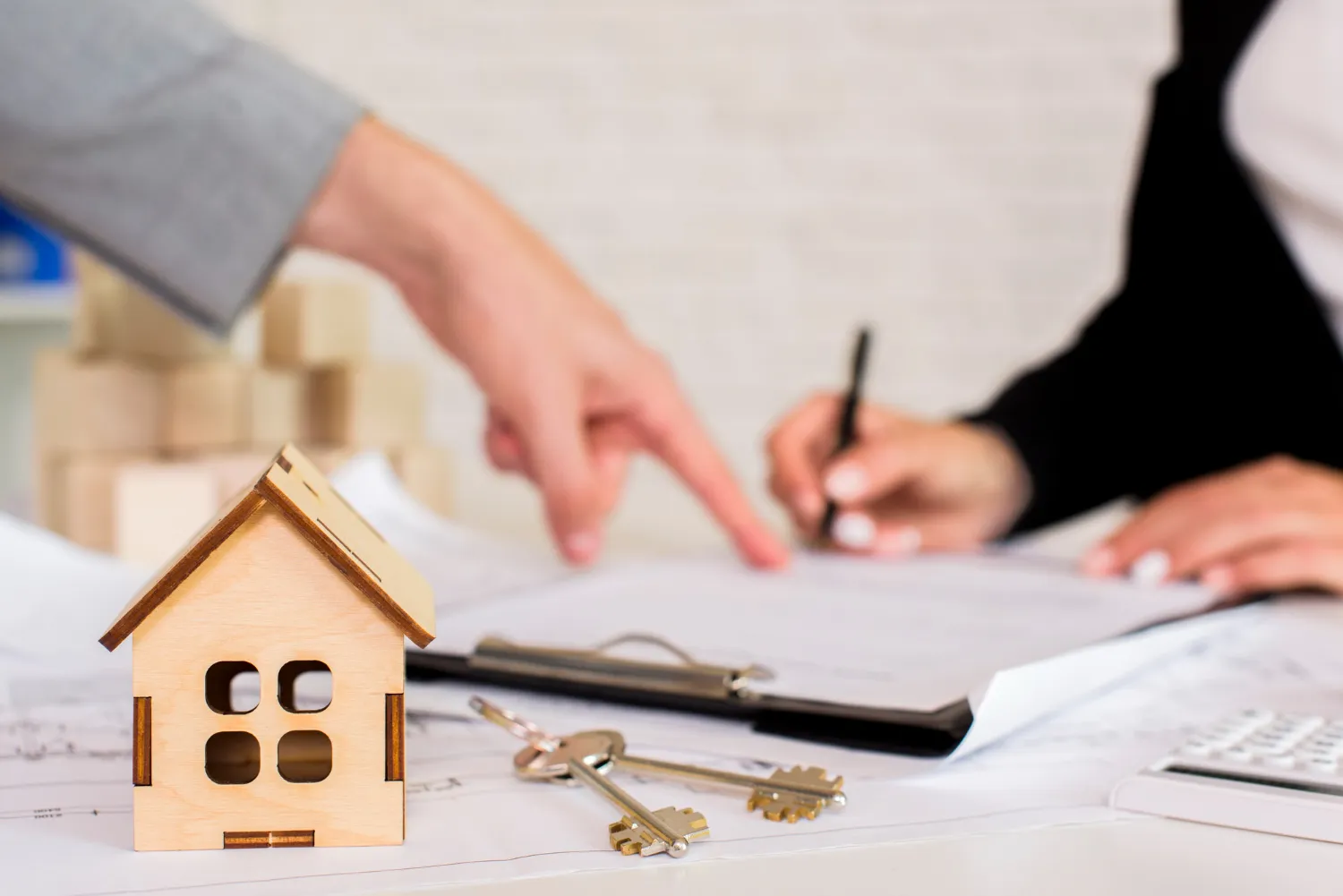 A miniature wooden house near keys and documents, as a hand gestures to a contract being signed by another person A miniature wooden house near keys and documents, as a hand gestures to a contract being signed by another person